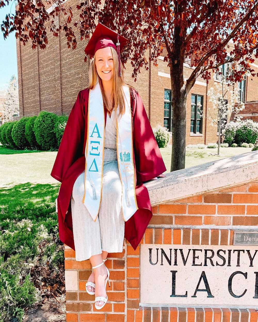 An Alpha Xi Delta woman poses in front of a university building wearing her cap and gown, and sorority stole.