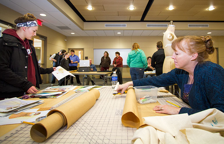 Arts Costuming Shop Manager Michelle Collyar working at a desk with students in the background.