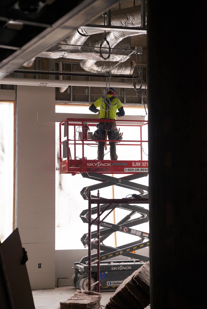 A worker installs drywall using a lift in front of one of the many large windows.