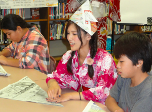 Image of Ayako Tsujita teaching origami to twoLogan Middle School students while seated at a table. 