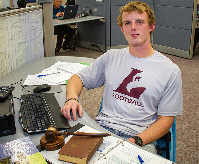 Image of Josh Gran sitting at a desk with computer in Murphy Library. A gavel is on the desk.