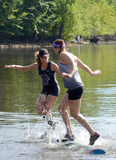 Image of Katie Rick on a log balancing with Emilhy Christopherson. Pictured on a lake.