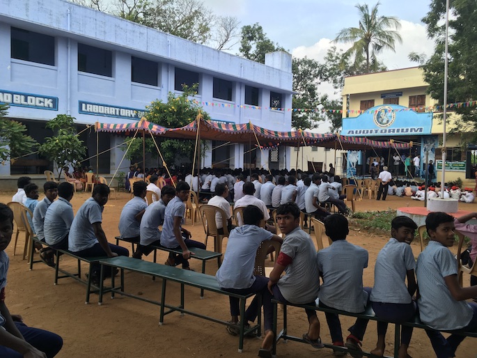 People seated in a courtyard of the school while Sondra LeGrand and others stand on stage. 