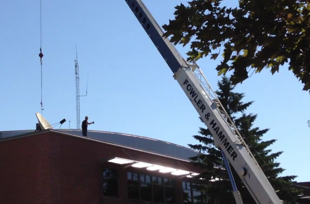 Image of man ontop of Cartwright Center giving a thumbs up as he stands near a satellite dish being removed from the roof. 