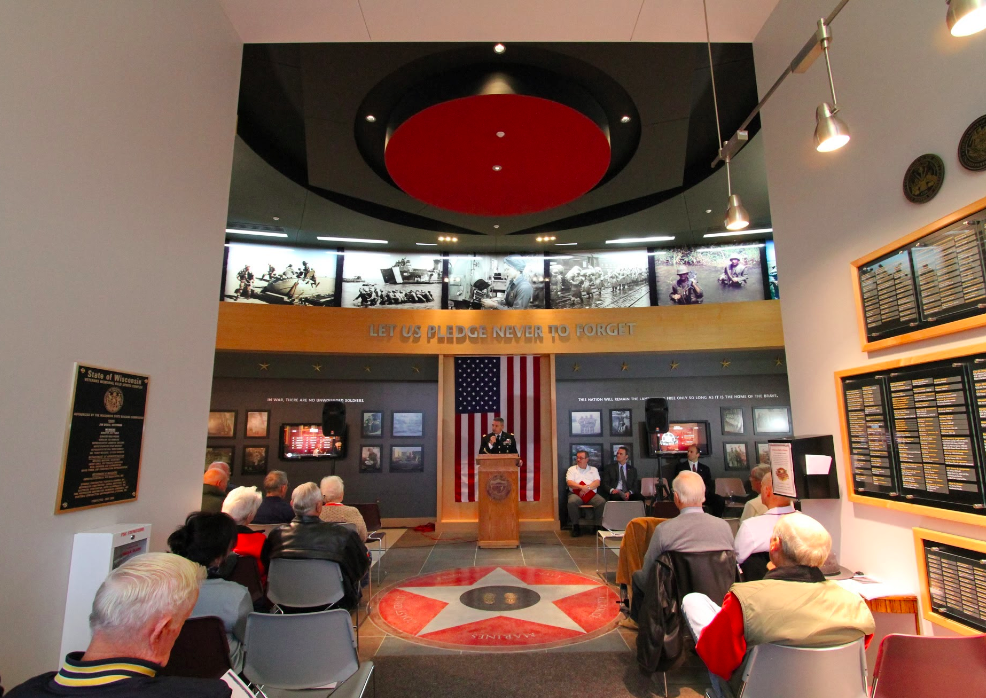 Image of people sitting in chairs and someone talking up front behind the podium in the Hall of Honor. 