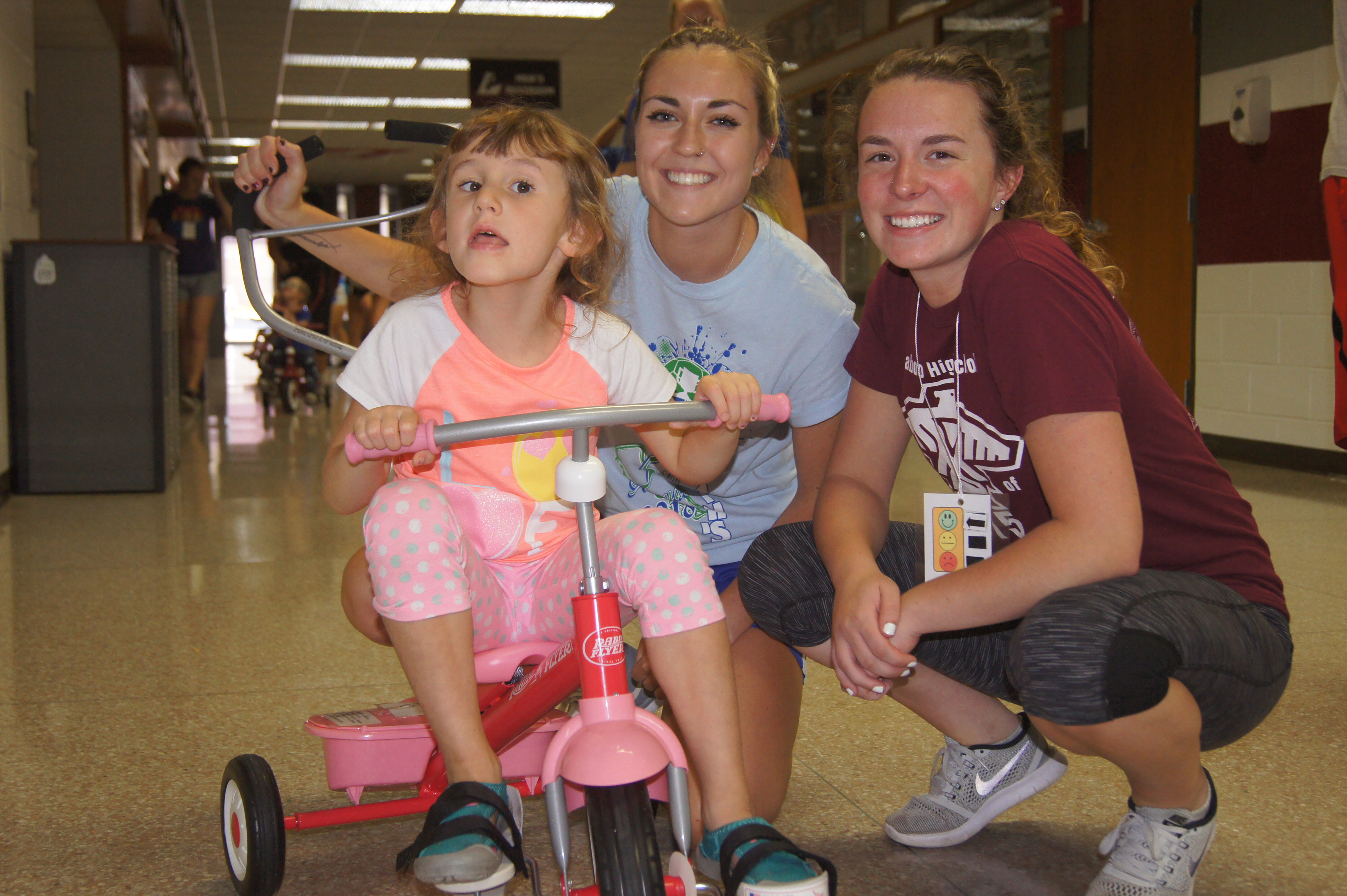 Two Adapted Physical Education Teaching Minors help a girl on a tricycle.